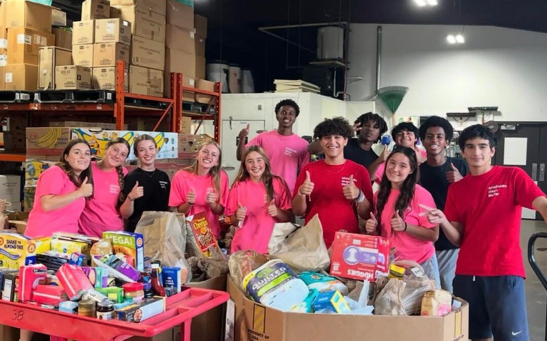 Group of people in pink/red shirts with food donations, giving thumbs up.