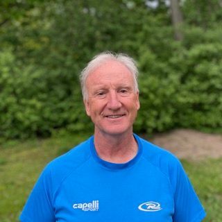Man in blue shirt smiles, standing outdoors in front of trees.