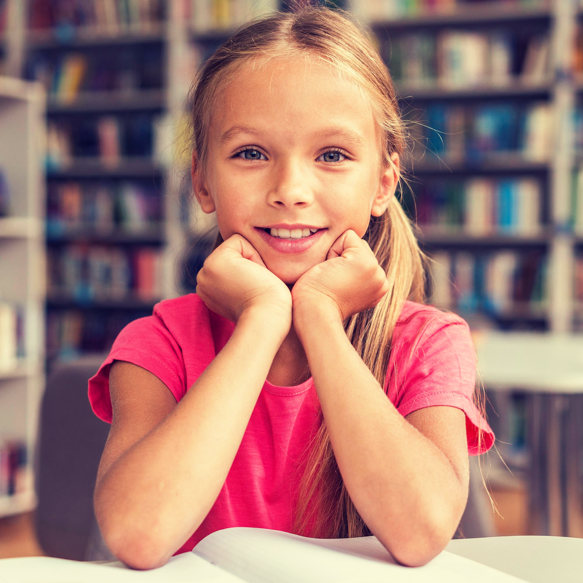 Een lachend kind in een roze shirt rust met haar kin op haar handen aan een tafel in de bibliotheek.