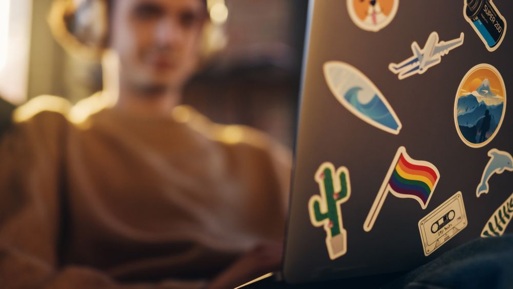 A Man is Sitting on a Couch Using a Laptop Computer With Stickers on It — Aldine Printers in Bungalow, QLD