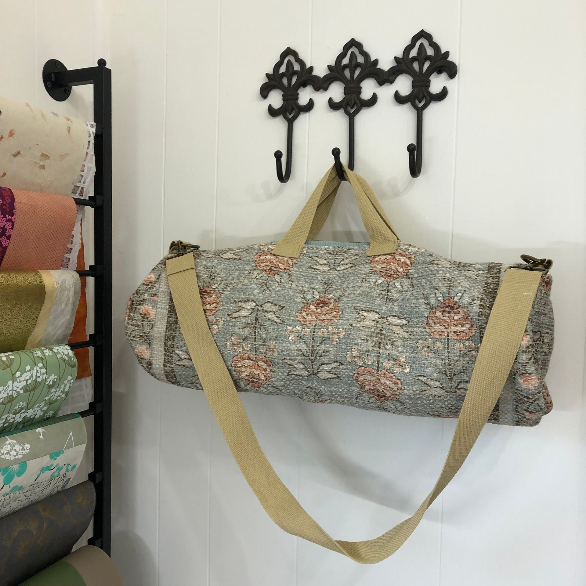 Floral patterned duffel bag hanging on a wall with ornate hooks, next to fabric rolls.