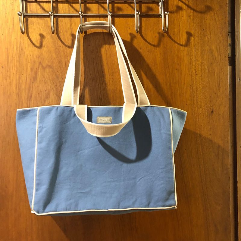A light blue canvas tote bag with white trim and beige handles hanging on a hook against a wooden door.