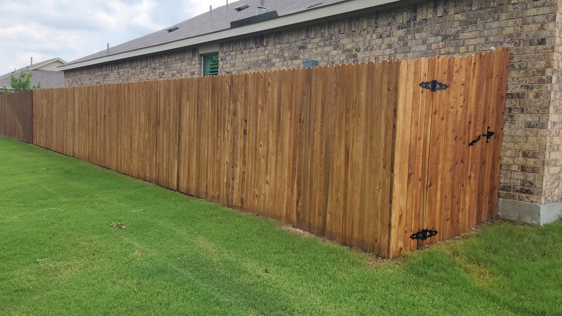 Wooden privacy fence with gate beside a stone house, bordering a green lawn