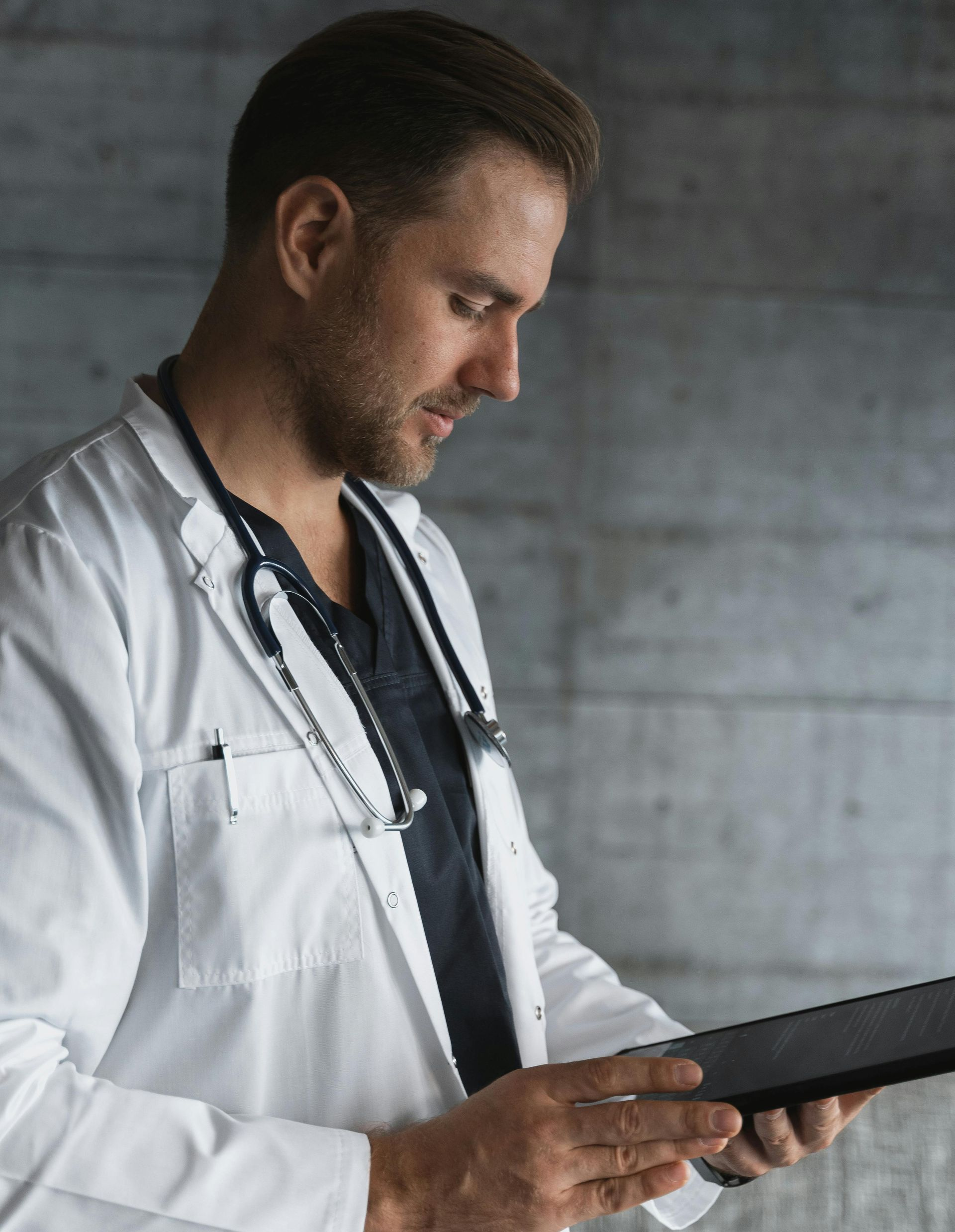 Doctor in white coat looking at tablet, stethoscope around neck, indoors.