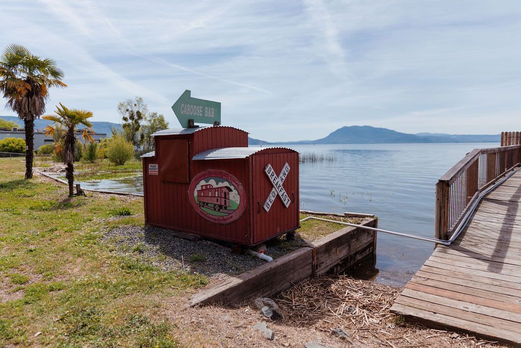 Rusty red container by a lakeside dock with a green sign, palm trees, and distant hills under a cloudy sky