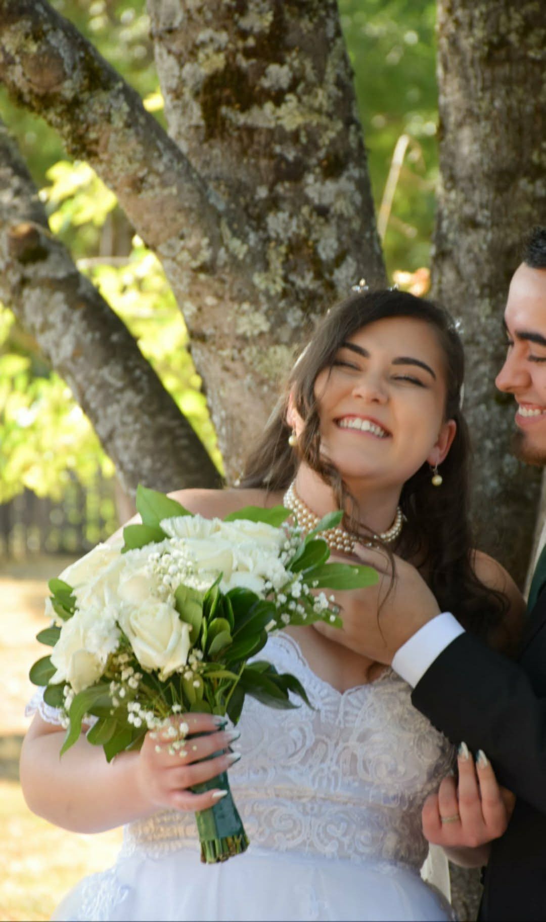 Smiling bride holding a white bouquet outdoors beside a man in a suit under a tree
