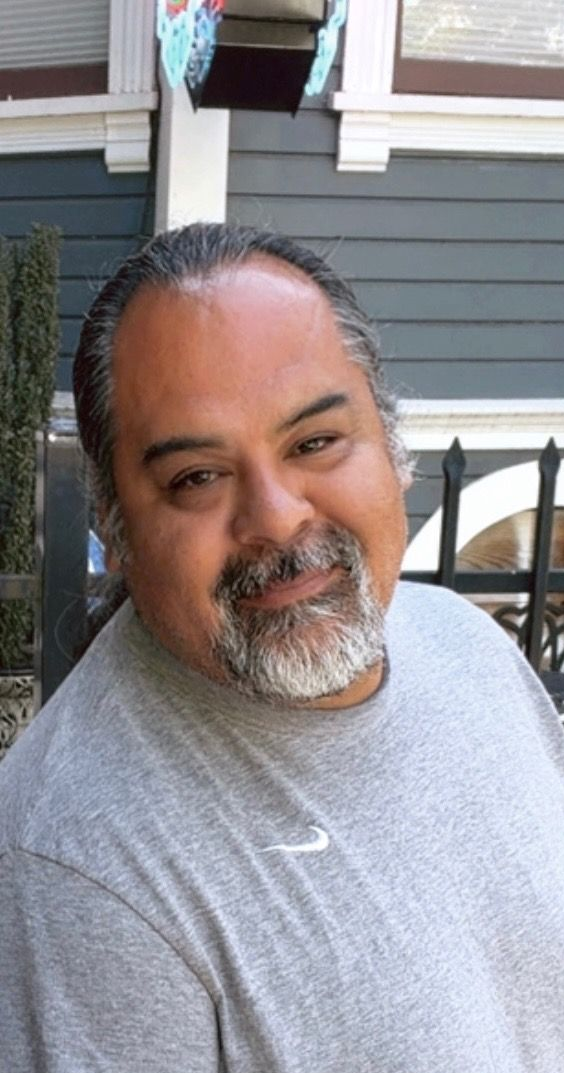 Smiling man in a gray shirt standing outdoors in front of a house