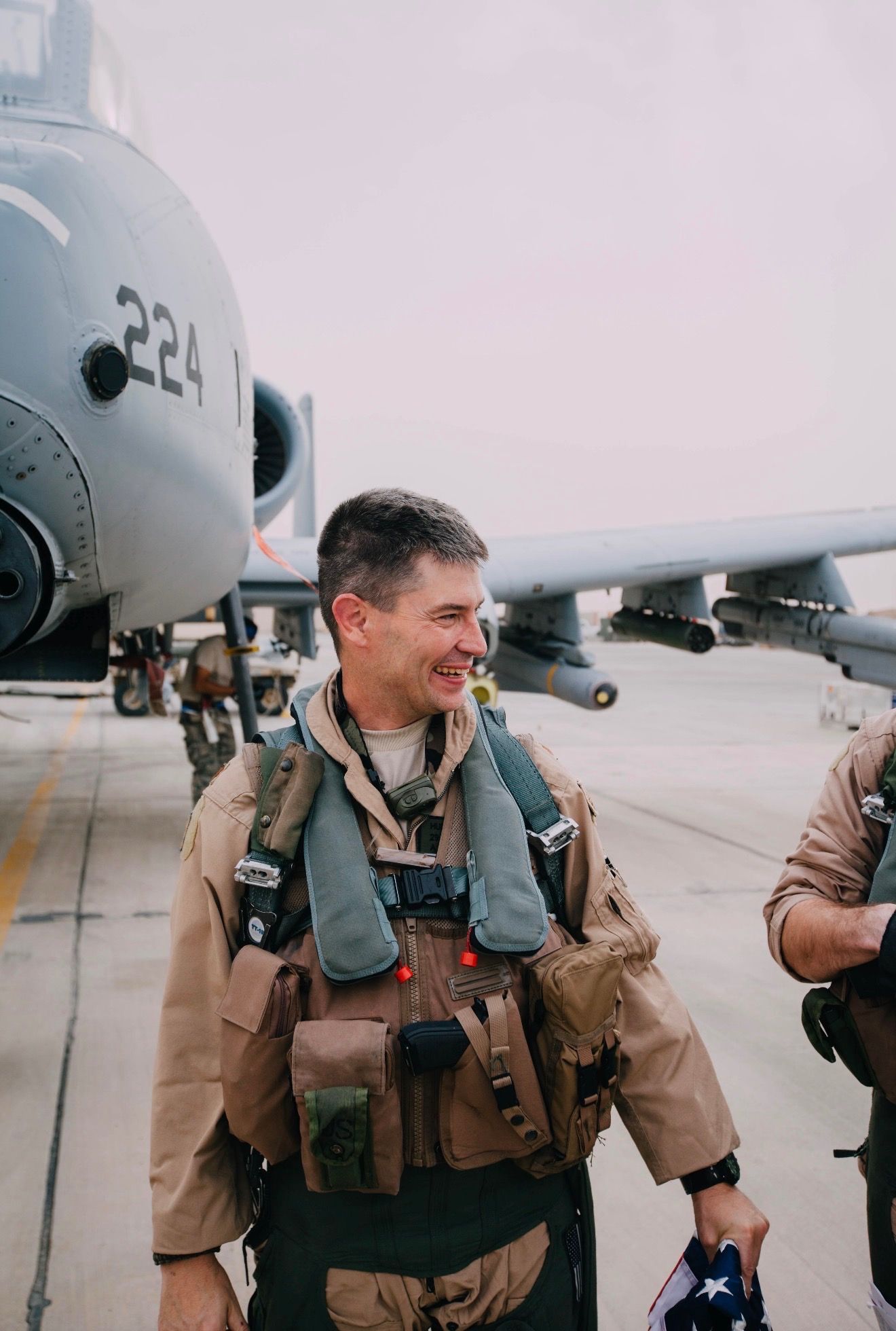 Smiling airman in tan flight gear stands beside a military aircraft on a tarmac.