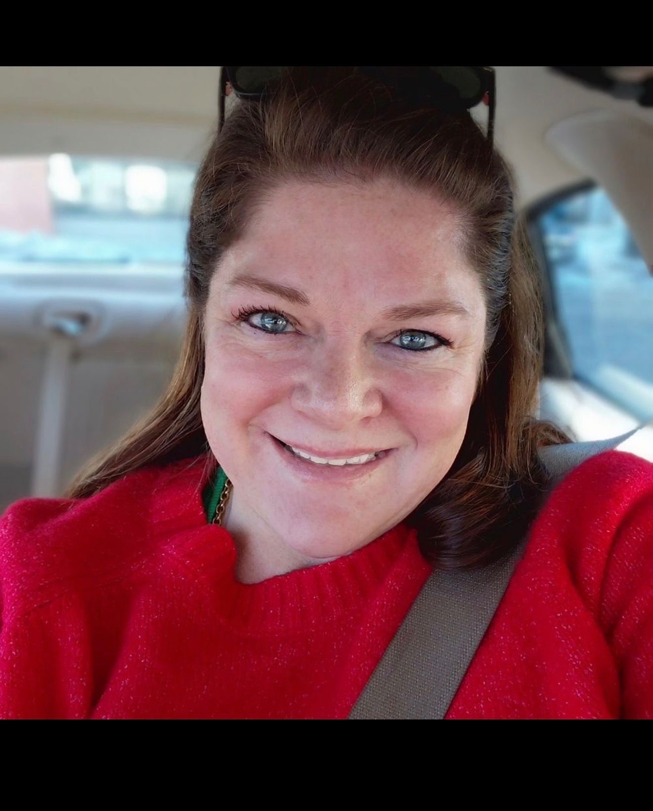 Smiling person in a red sweater sitting in a car seat, wearing a seatbelt.