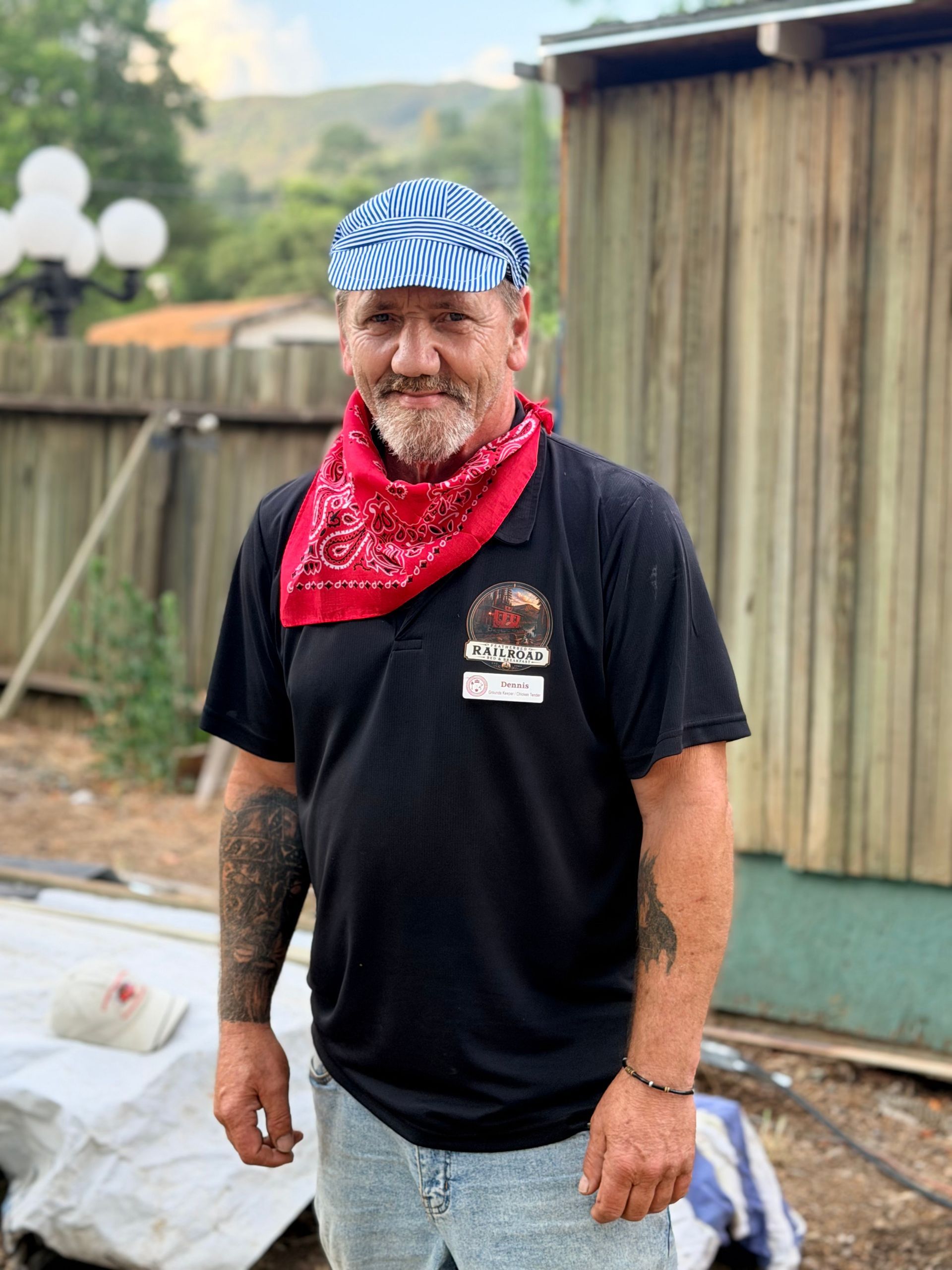 Man in a blue striped cap and red bandana standing in a backyard near a wooden fence