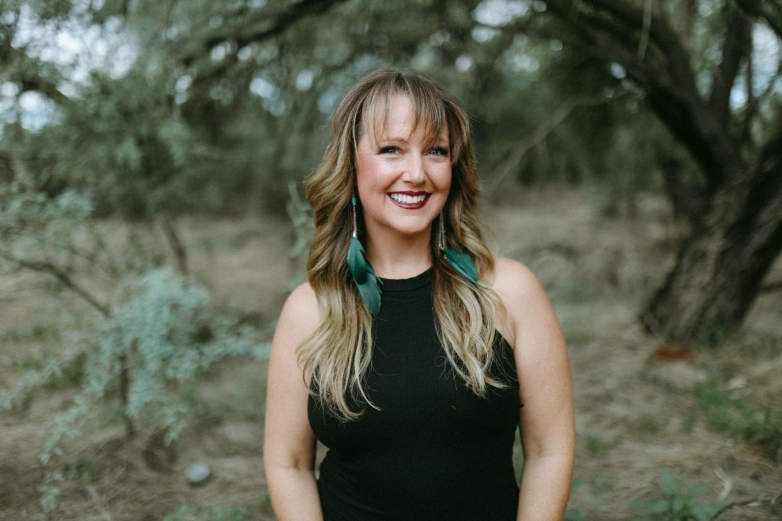 Smiling woman in a black sleeveless top standing outdoors among trees and greenery