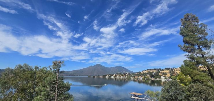 Mountain lake with a calm reflection, blue sky and clouds, framed by trees and distant hills