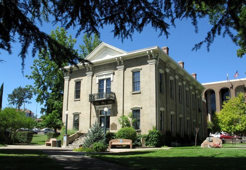 Historic stone building with columns and a balcony, framed by trees on a sunny campus lawn.