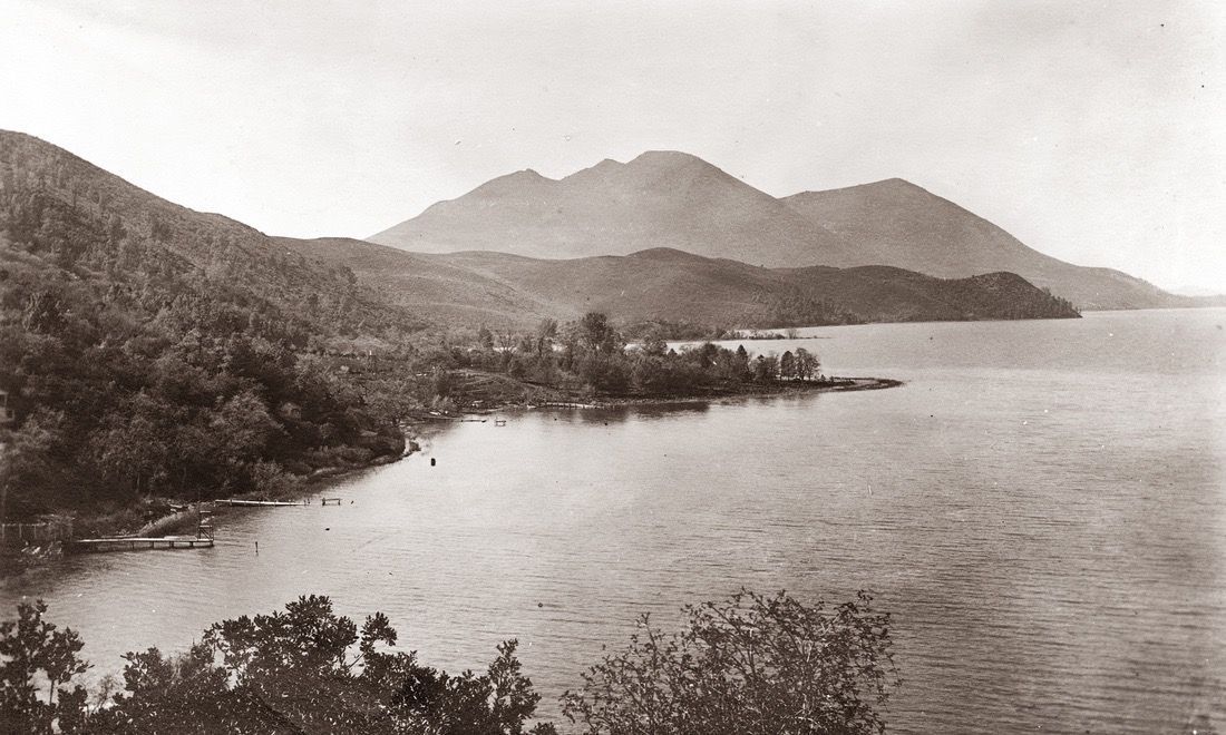 Sepia-toned coastal landscape with mountains, wooded shoreline, and calm bay water