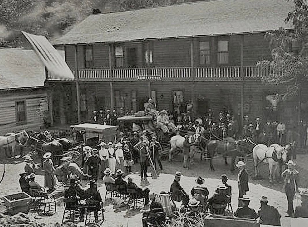 Crowded outdoor gathering with horses and carriages in front of a long two-story building