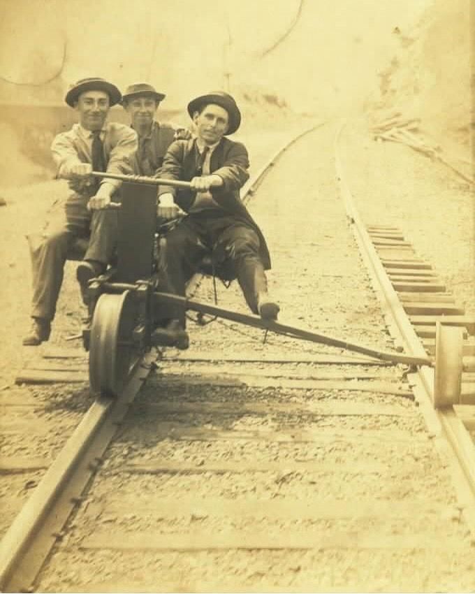 Three men ride a rail handcar on wooden tracks in a sepia-toned historical photo.