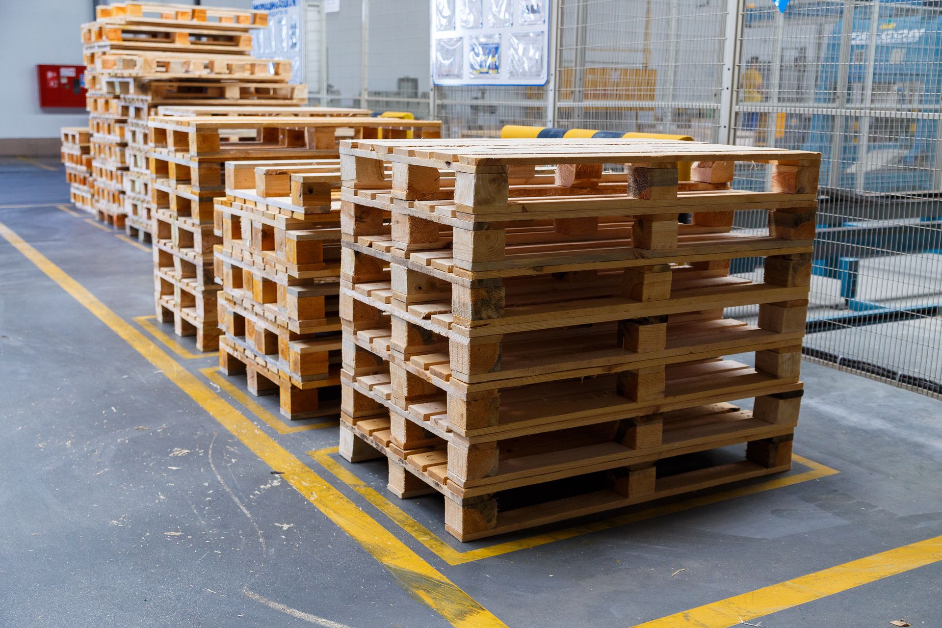 Wooden pallets stacked in an industrial setting, next to yellow painted lines on the floor.