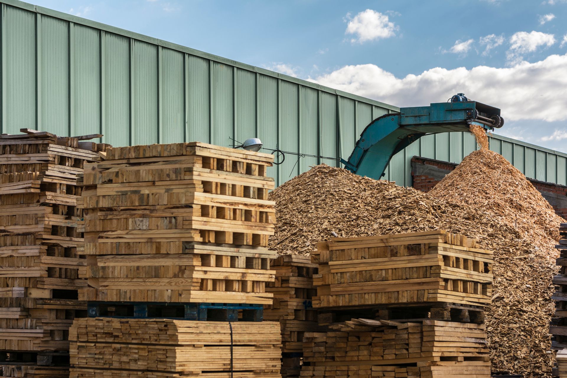 Wooden pallets stacked near wood chipper, processing wood chips. Factory setting with blue sky.