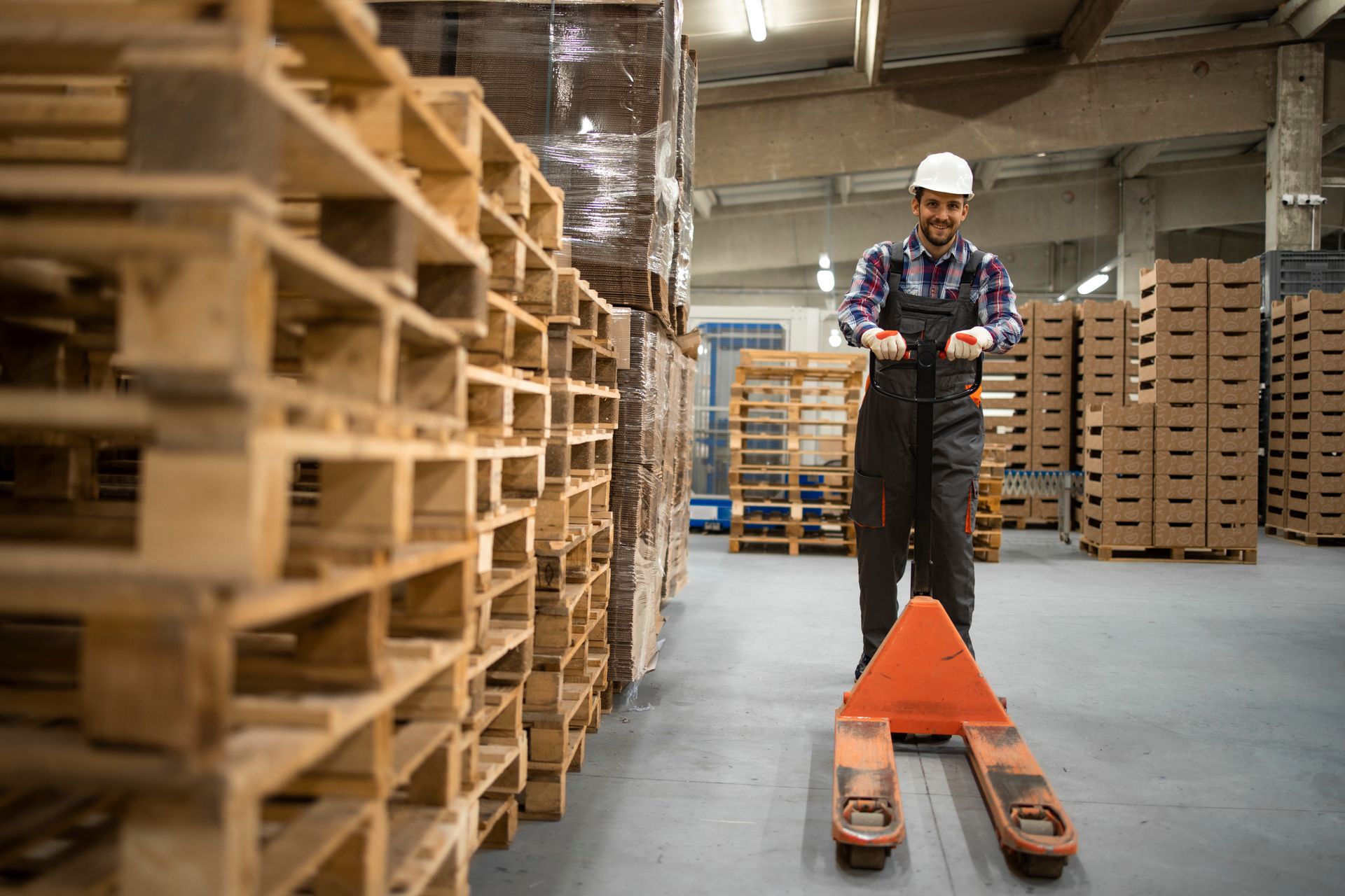 Warehouse worker operating a pallet jack near stacks of wooden pallets.