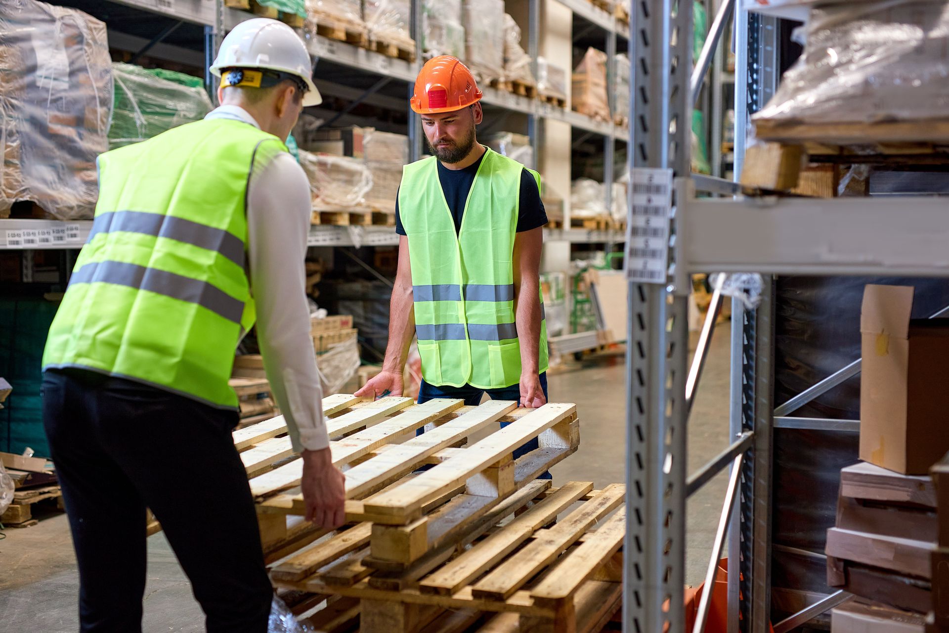 Two workers in safety vests and helmets lifting wooden pallets in a warehouse.