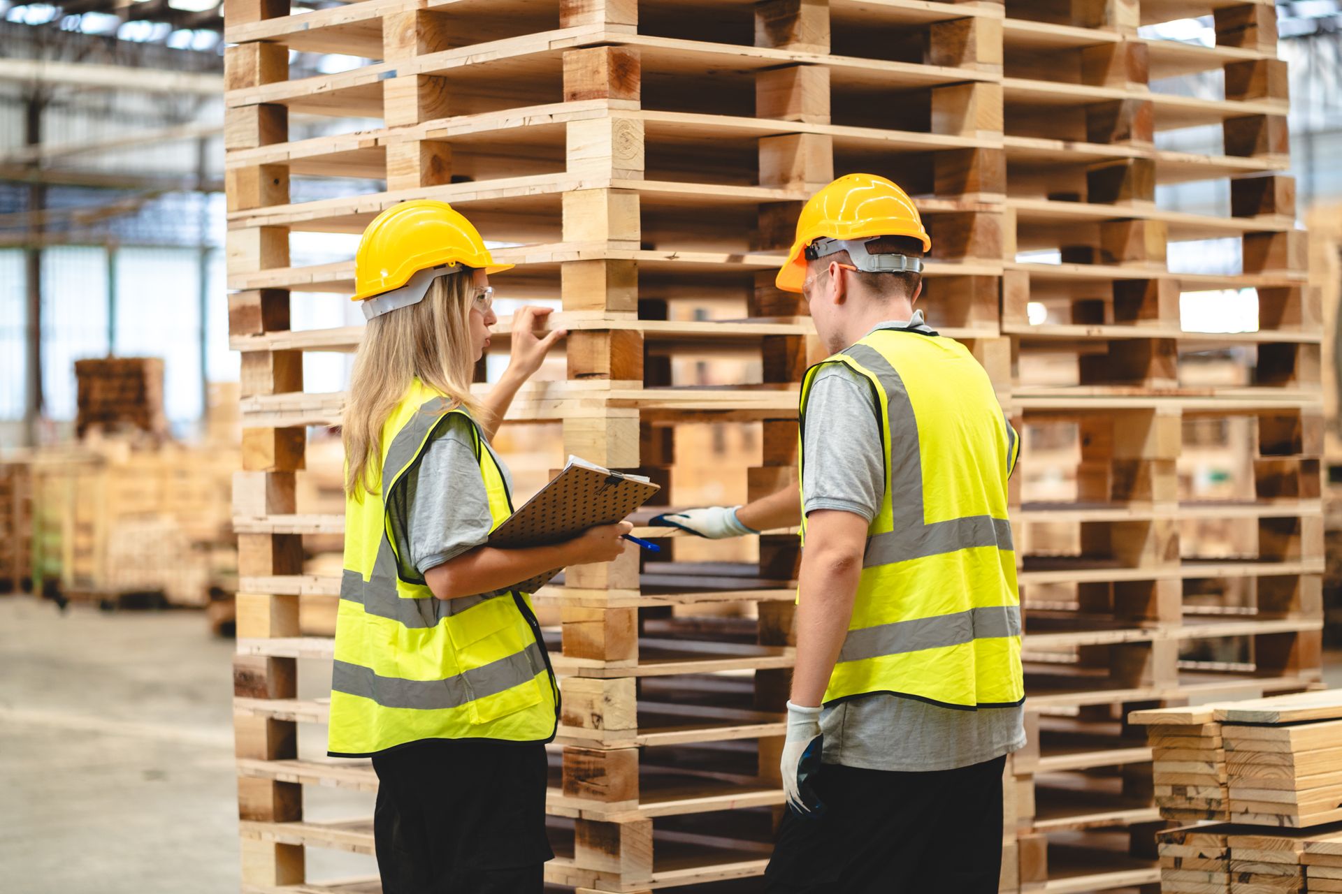 Two people in safety vests and hard hats inspecting stacked wooden pallets in a warehouse.