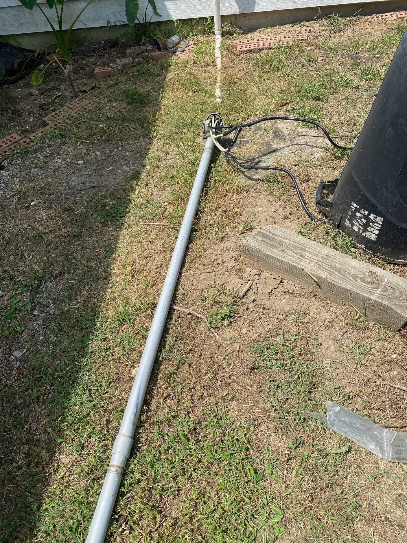 Gray pipe on grass beside a black utility pole and concrete blocks, with a cable lying nearby.