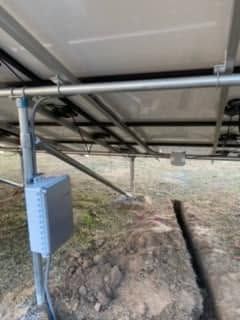 Underside of a solar panel array with metal supports beside a dirt trench outdoors