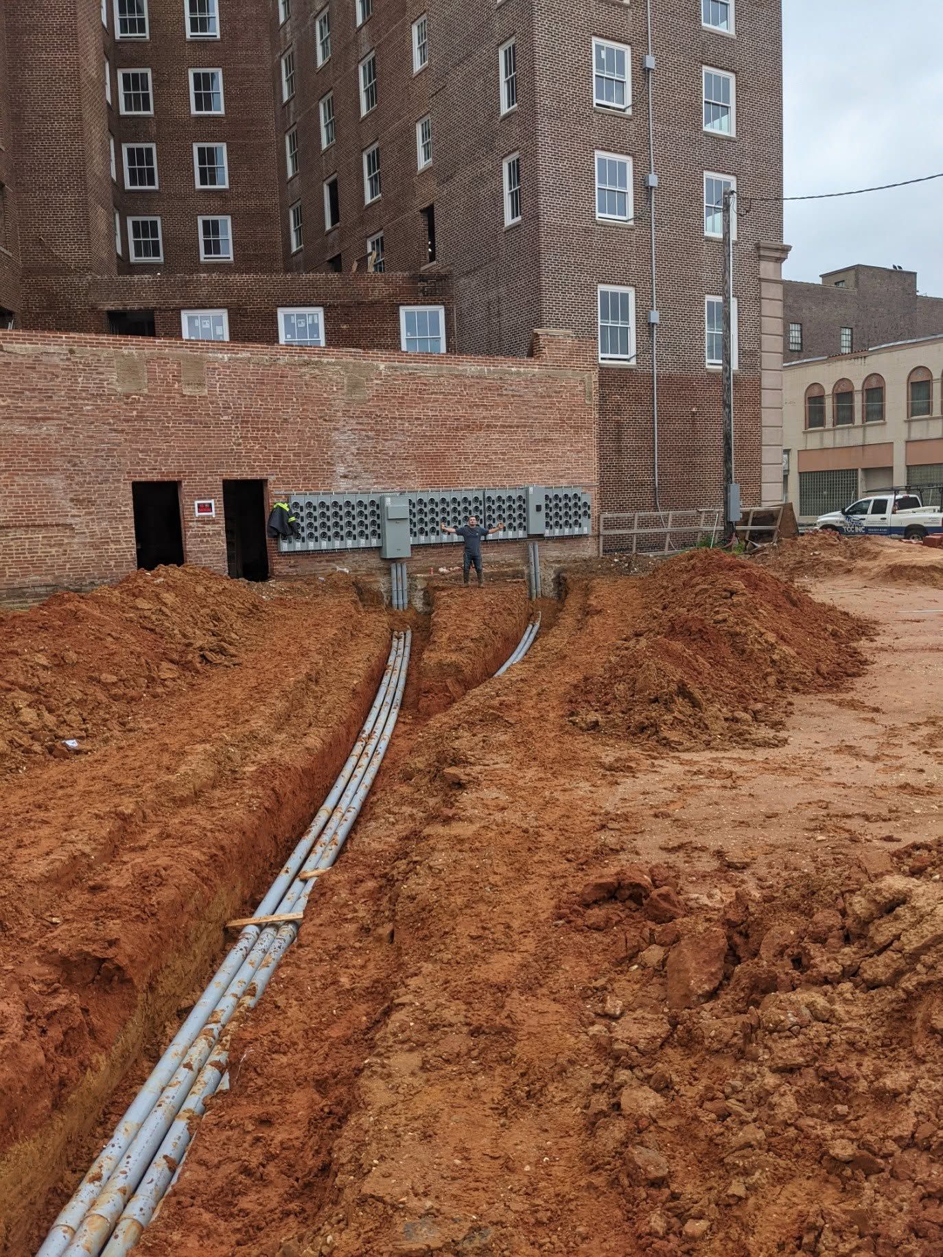 Construction site with gray utility conduits running through a red dirt trench beside a brick building