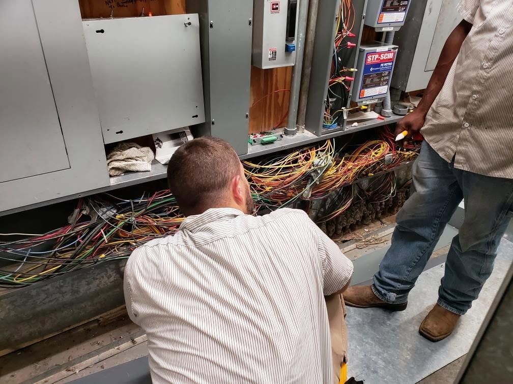 Technicians inspecting exposed wiring and electrical equipment inside a cabinet room.
