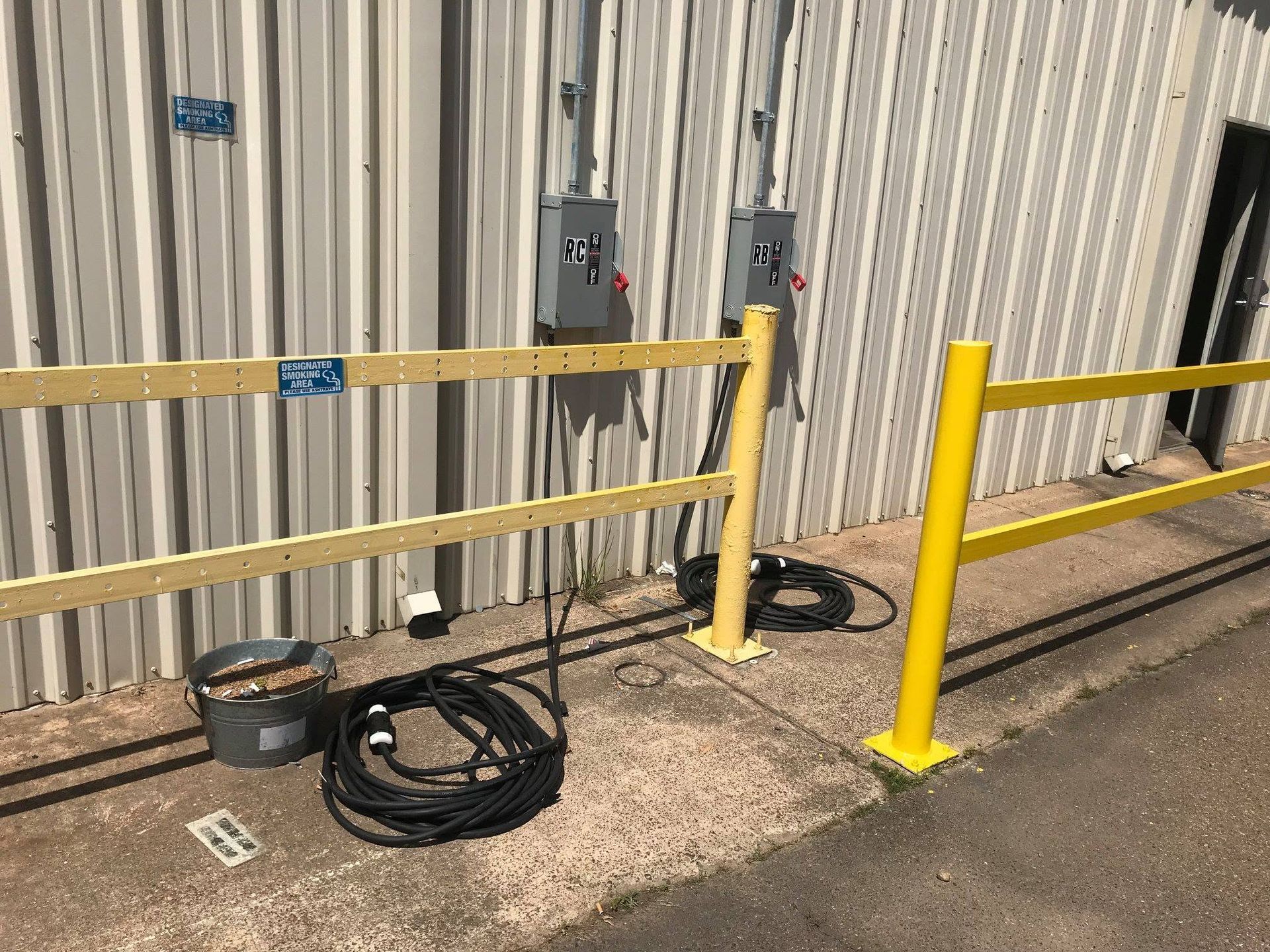 Yellow safety barrier and bollards in front of a corrugated metal wall with electrical boxes and coiled cables.