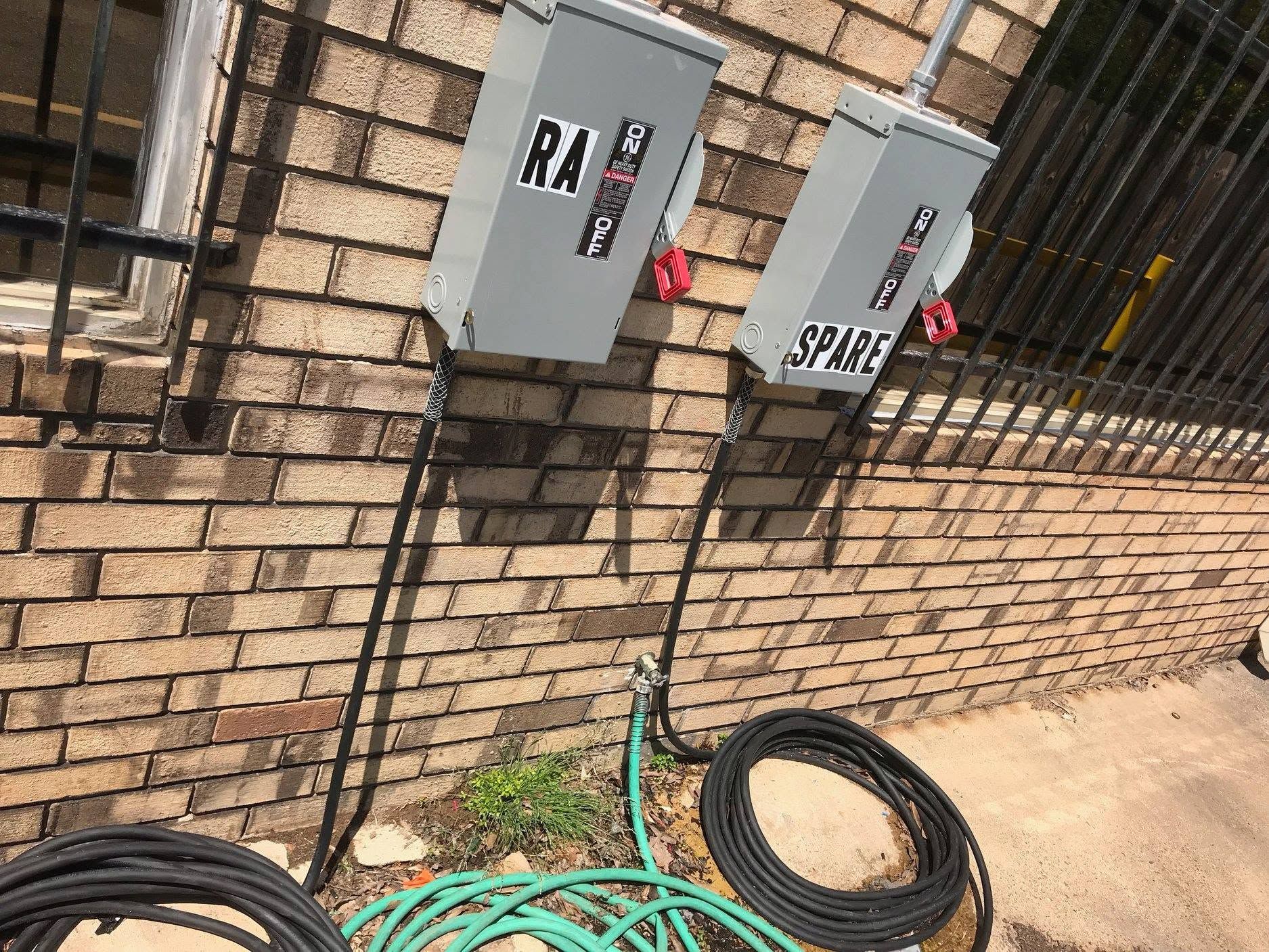 Two gray electrical boxes on a brick wall with coiled cables on the ground