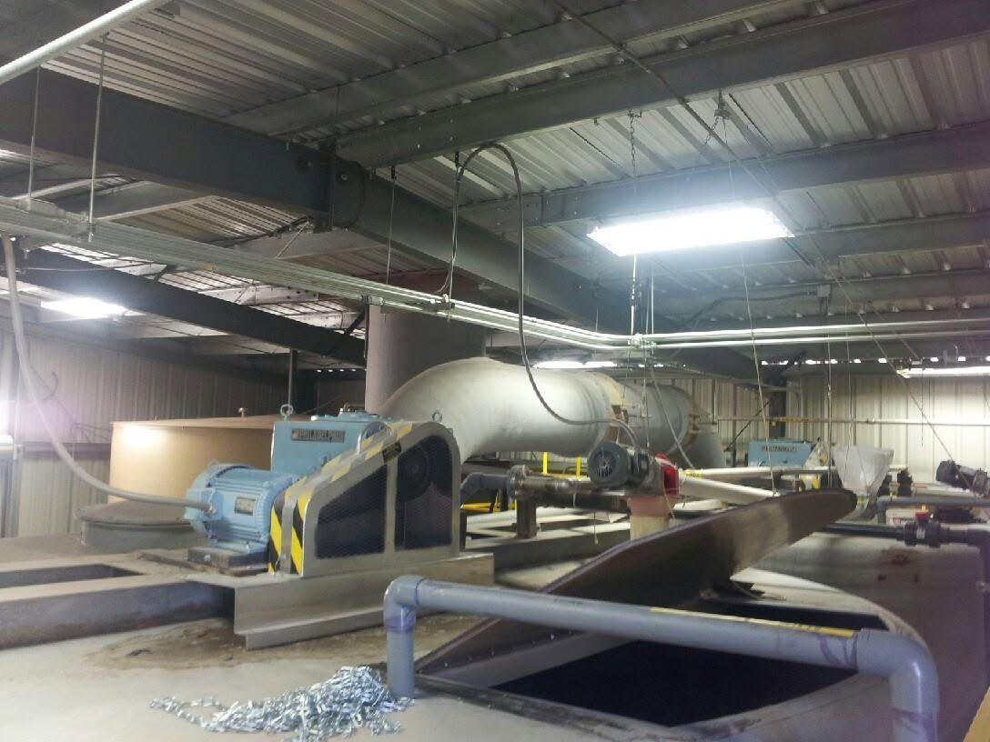 Industrial laundry room with large machines, pipes, and workers under bright overhead lights