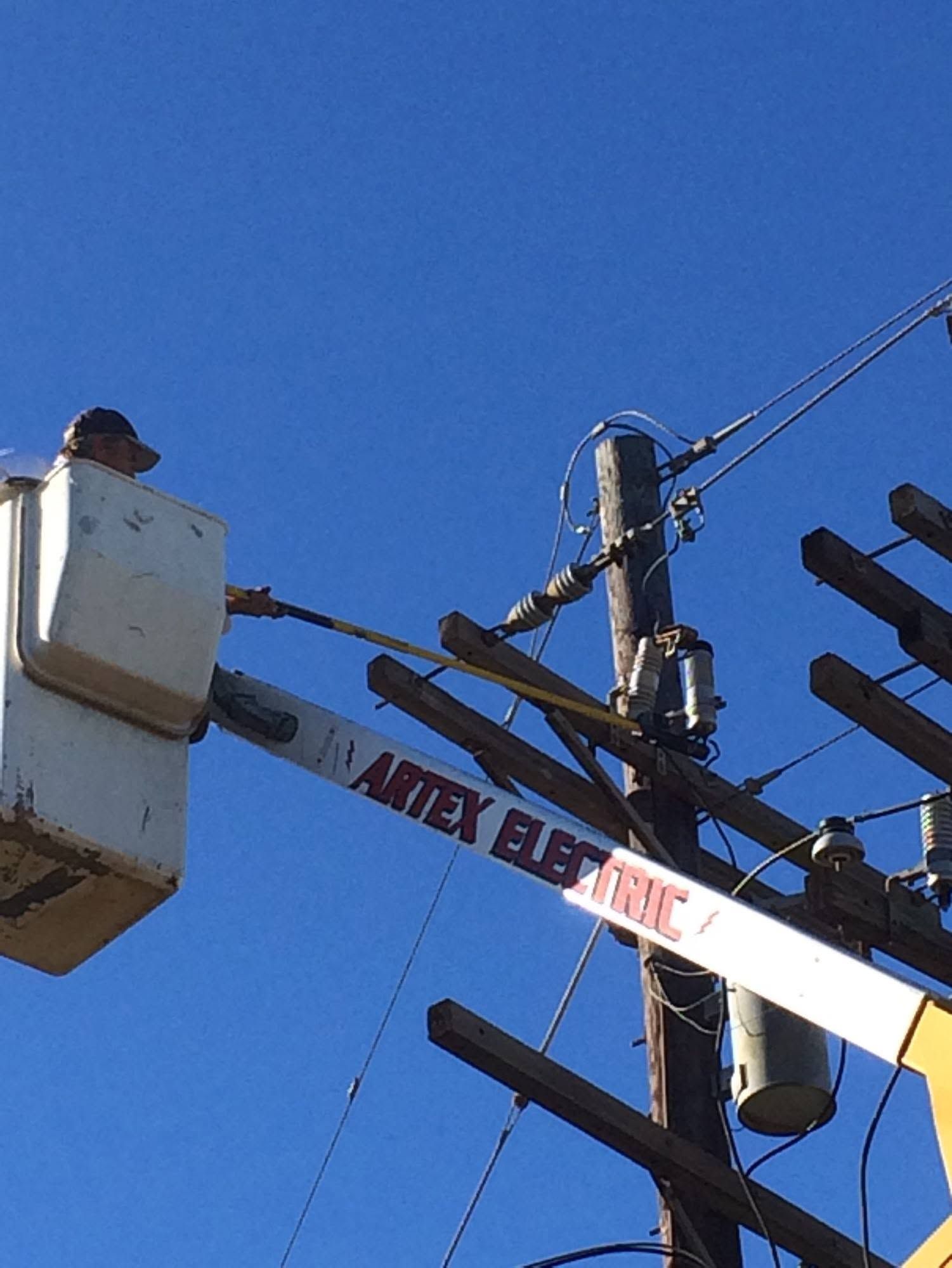 Utility worker in a bucket lift repairing overhead power lines against a clear blue sky