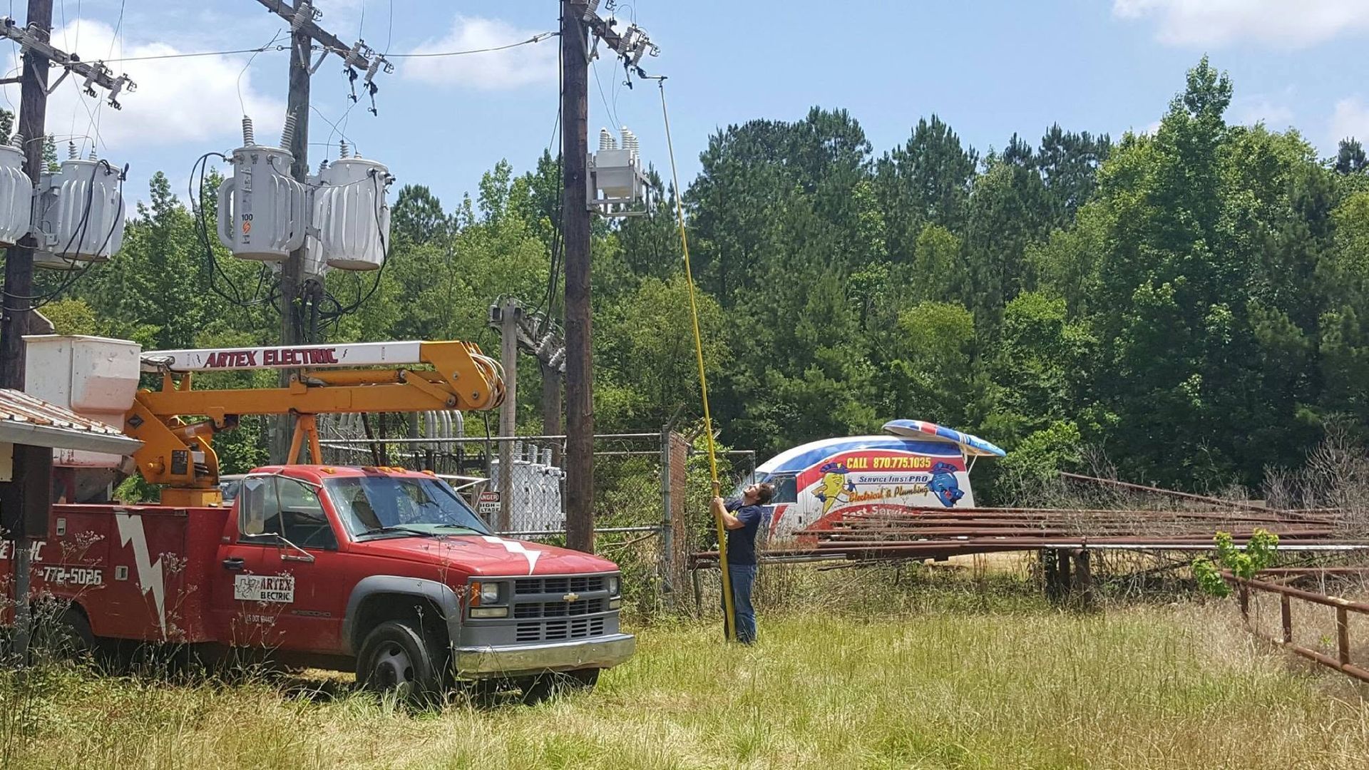 Red utility truck with bucket lift near workers and stacked wood in a grassy clearing by trees