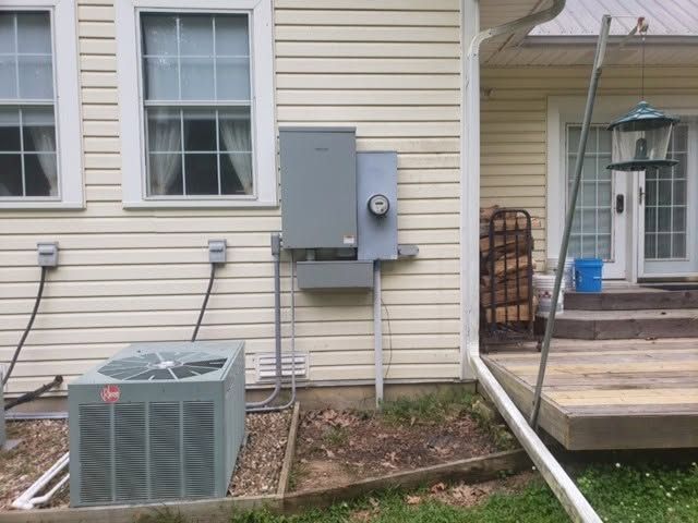 Outdoor HVAC unit beside beige house siding and porch, with electrical panel and blue bucket nearby