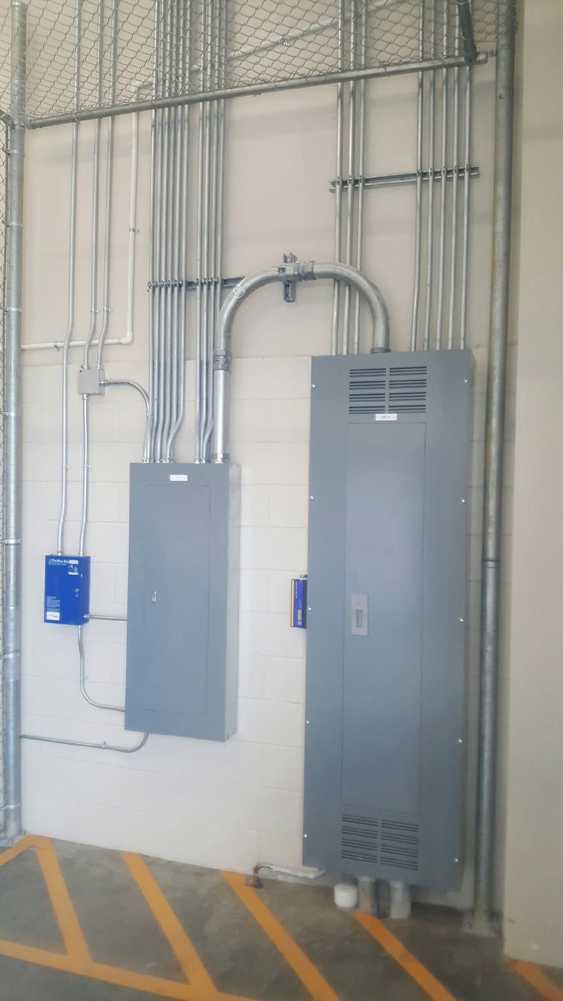 Electrical panels and conduit mounted on a wall in a utility area with yellow floor markings.