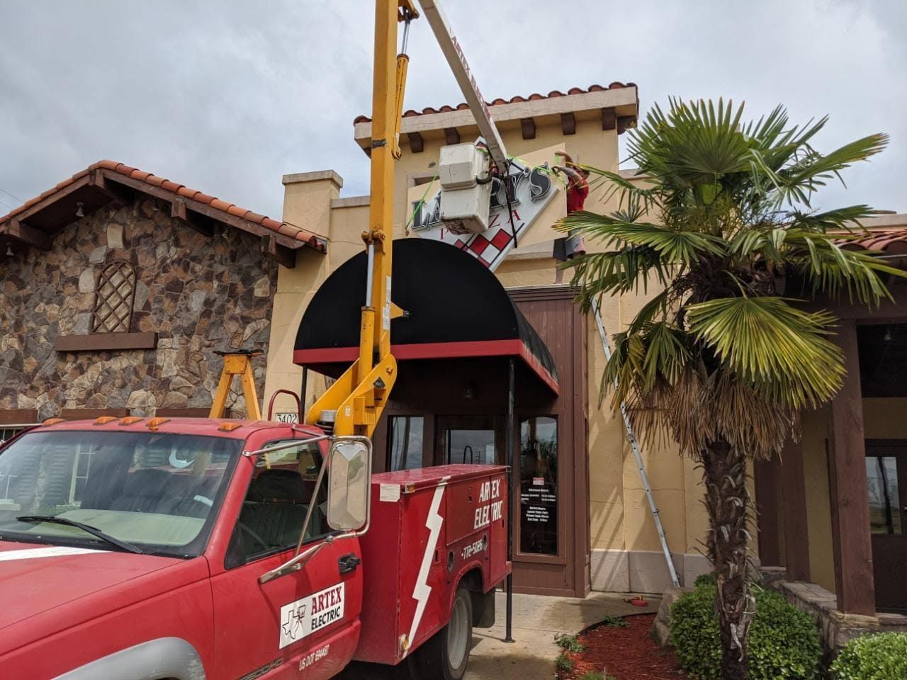 Red utility truck with crane parked beside a stucco storefront and palm tree.