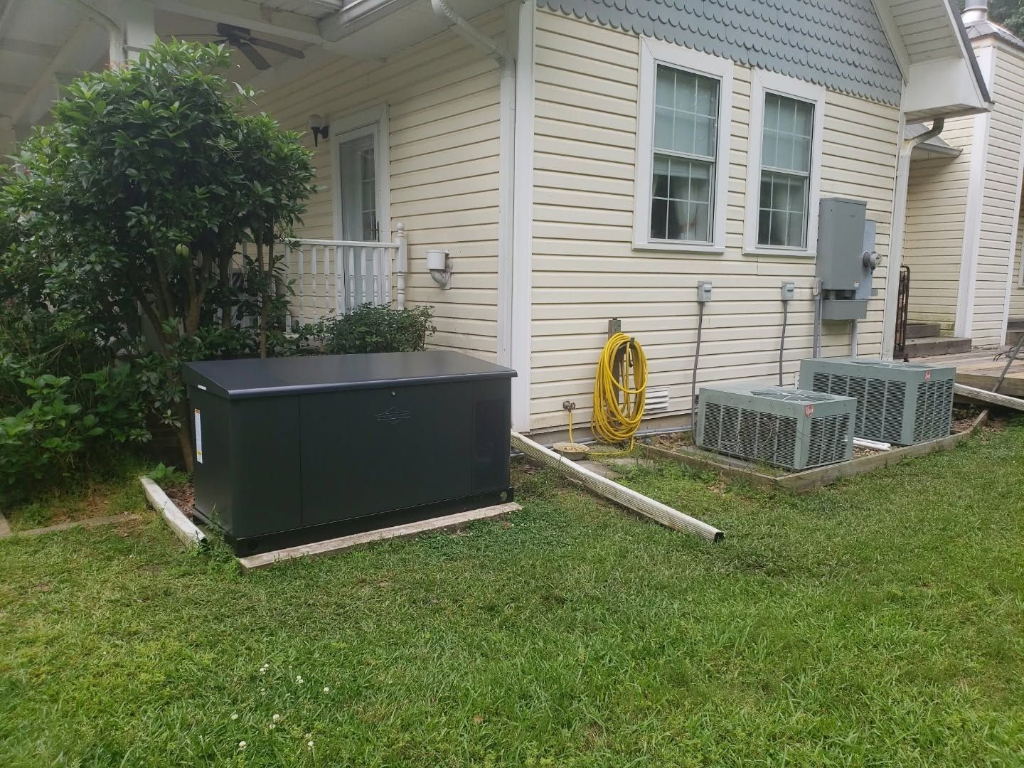 Side yard of a house with an air conditioner unit, garden hose, and outdoor utility boxes on grass.