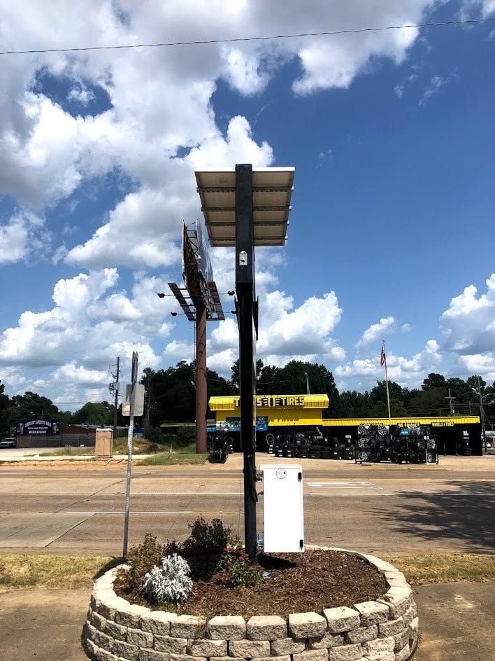 Solar-powered signpost on a stone planter by a road, with a yellow bus and cloudy blue sky behind it