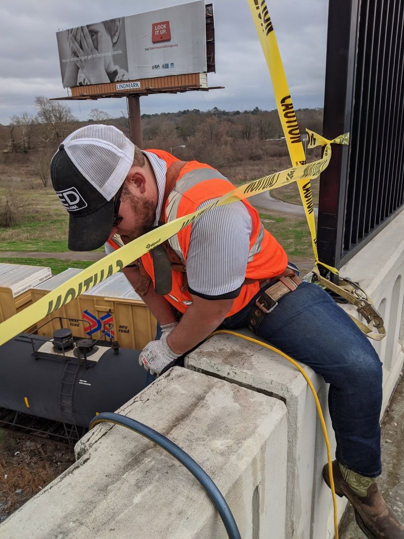 Worker in orange vest and hard hat crouches on a concrete barrier at a taped-off roadside worksite.