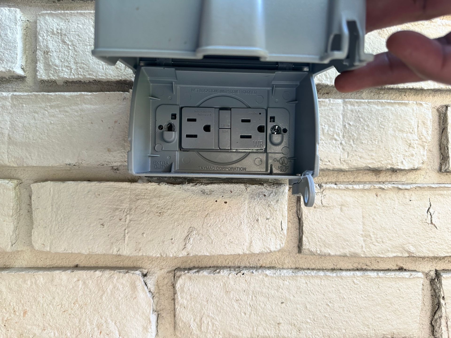 Open gray outdoor electrical outlet box mounted on a brick wall, with a hand touching the cover.