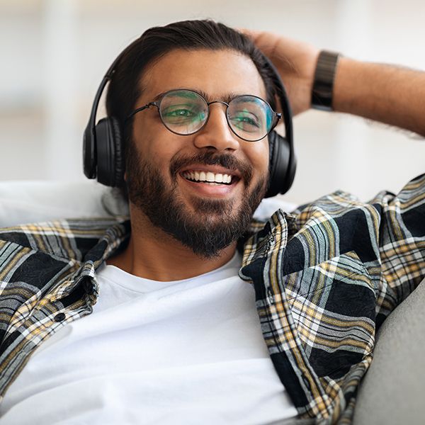 A man wearing headphones and glasses is smiling while sitting on a couch.