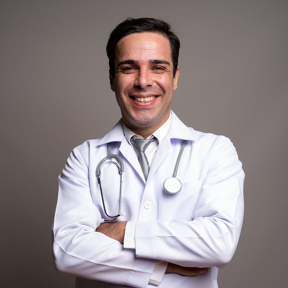 A smiling doctor with a stethoscope around his neck is standing with his arms crossed.