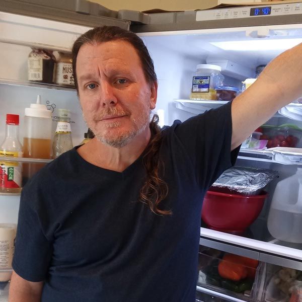 Man standing in an open refrigerator, wearing a dark shirt and looking at the camera.