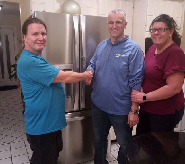 Three people smiling and shaking hands in a  room beside a stainless-steel refrigerator.