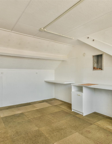 Empty white room with sloped ceiling, built-in desk, and beige carpet flooring.