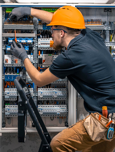 Technician in orange hard hat adjusts wiring in an open electrical control panel.