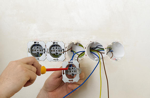 Hand wiring electrical outlets in a wall, with a screwdriver and exposed colored cables.