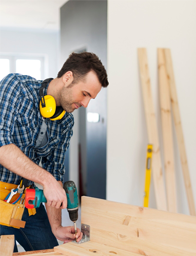 Man using a drill on a wooden board in a home workshop, wearing yellow ear protection and a tool belt