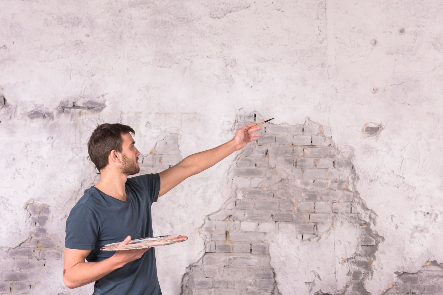 Man holding a putty knife and pointing at a damaged plaster wall with exposed bricks